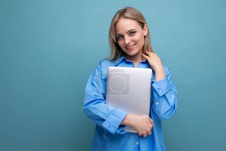 Téléchargez les photos : Timide fille blonde souriante étudiante avec un ordinateur portable sur un fond bleu vif. - en image libre de droit