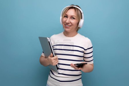 Téléchargez les photos : Portrait de mignon jeune étudiante blonde souriante avec de gros écouteurs sans fil et ordinateur portable sur fond bleu avec espace de copie. - en image libre de droit
