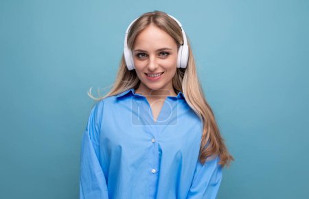 Téléchargez les photos : Fille blonde souriante en chemise décontractée écoutant podcast dans de gros écouteurs. - en image libre de droit