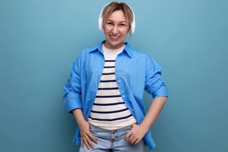 Foto de Chica rubia sonriente caucásica vestida con un suéter a rayas y una camisa casual escucha música en la aplicación en auriculares inalámbricos sobre un fondo azul. - Imagen libre de derechos