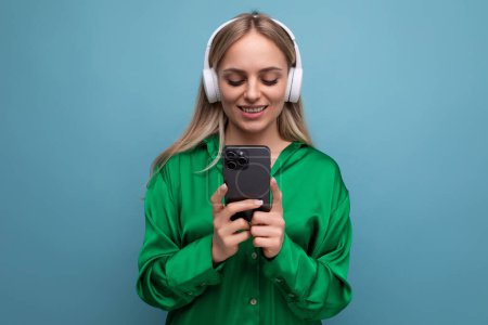 Foto de Foto de una linda joven rubia atractiva en auriculares con un teléfono inteligente en sus manos sobre un fondo azul. - Imagen libre de derechos