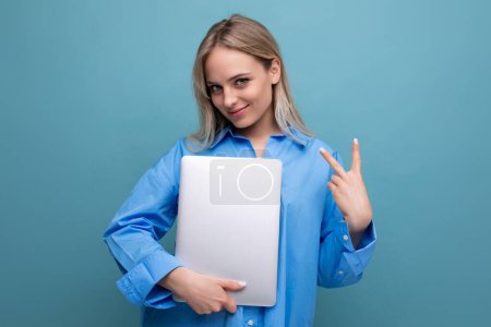 Téléchargez les photos : Mignon fille blonde étudiant avec un ordinateur portable sur un fond bleu vif. - en image libre de droit