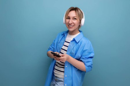 Foto de Mujer rubia adorable europea en camisa escuchando música en auriculares inalámbricos sobre fondo azul. - Imagen libre de derechos
