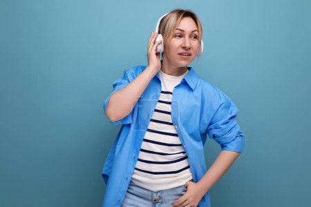 Téléchargez les photos : Fille blonde lumineuse vêtue d'une chemise décontractée écoute de la musique dans l'application dans les écouteurs sans fil sur un fond bleu. - en image libre de droit