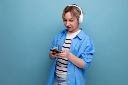 Foto de Chica rubia brillante vestida con una camisa casual escucha música en la aplicación en auriculares inalámbricos sobre un fondo azul. - Imagen libre de derechos