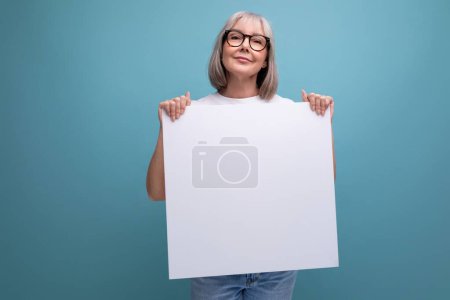 60s mature retired woman with gray hair holding blank billboard with mockup on studio background.
