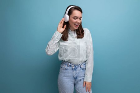 Téléchargez les photos : Énergique jolie brune mignonne jeune femme en chemise écoutant de la musique sur fond studio avec rythme de copie. - en image libre de droit