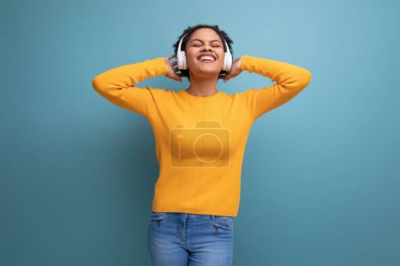 Foto de Mujer latina bastante positive20s con pelo afro en suéter amarillo casual escuchando música en auriculares inalámbricos. - Imagen libre de derechos