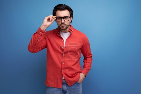 Téléchargez les photos : Surpris caucasien brunette 30s homme dans une chemise rouge tenant des lunettes en urki sur fond avec espace de copie. - en image libre de droit