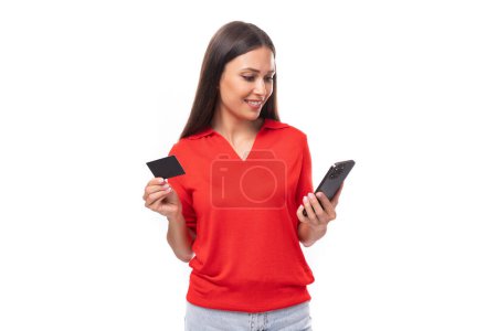 Téléchargez les photos : Jeune jolie femme européenne aux cheveux noirs est habillée d'un t-shirt rouge avec un smartphone et une carte plastique sur un fond blanc. - en image libre de droit