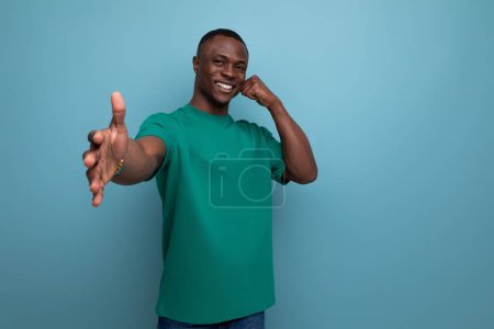 Téléchargez les photos : Jeune sympathique beau mec africain avec une coupe de cheveux courte habillé dans un t-shirt de base souriant sur le fond avec espace de copie. - en image libre de droit