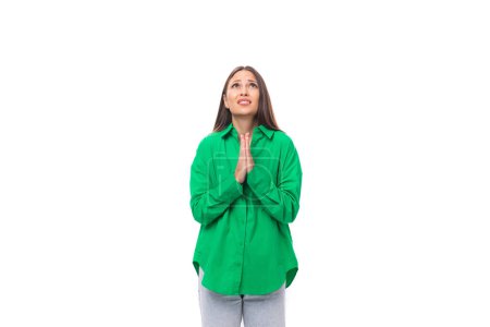 Foto de Mujer morena europea delgada rezando con el pelo largo y los ojos marrones vestidos con la camisa verde casual esperanzas. - Imagen libre de derechos