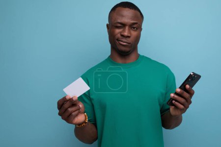 Foto de Un joven africano con un corte de pelo corto vestido con una camiseta básica utiliza un teléfono inteligente para transferencias desde una tarjeta bancaria en el fondo con espacio para copiar. - Imagen libre de derechos