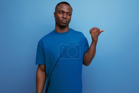 Foto de Joven hombre americano en una camiseta muestra su mano a un lado sobre un fondo azul con espacio para copiar. - Imagen libre de derechos