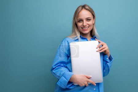 Téléchargez les photos : Sourire joyeux blond fille étudiante avec un ordinateur portable sur un fond bleu vif. - en image libre de droit