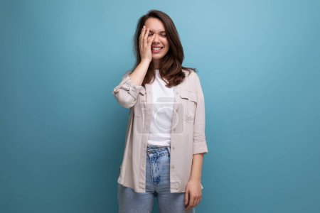 Téléchargez les photos : Modeste brune jeune femme adulte en chemise et jeans couvert son visage avec sa main sur un fond bleu. - en image libre de droit