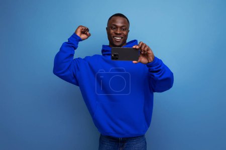 Téléchargez les photos : Positif jeune homme américain aux cheveux courts se réjouit tout en regardant la vidéo sur smartphone. - en image libre de droit