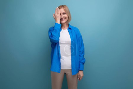 Téléchargez les photos : Modeste femme mature aux cheveux blonds dans une chemise décontractée sur un fond bleu avec espace de copie. - en image libre de droit