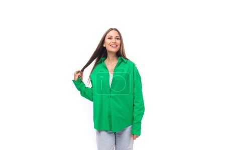Foto de Sonriente joven modelo femenina europea feliz con el pelo negro bien arreglado y maquillaje vestido con una camisa verde sobre un fondo blanco con espacio para copiar. - Imagen libre de derechos