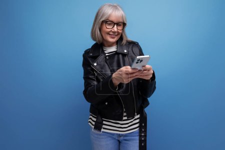 Photo for Smart 60s modern middle-aged woman grandmother with gray hair makes an order on the Internet using a smartphone on a bright background. - Royalty Free Image