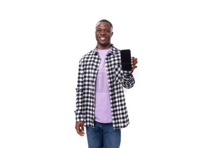 Foto de Un chico africano de 30 años vestido con una camisa a cuadros en blanco y negro demuestra la pantalla de un teléfono inteligente en el fondo con espacio para copiar. - Imagen libre de derechos