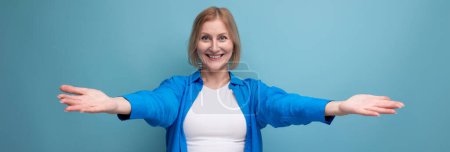 Téléchargez les photos : Femme mature aux cheveux blonds dans une chemise décontractée étend ses bras étreignant sur un fond bleu avec espace de copie. - en image libre de droit