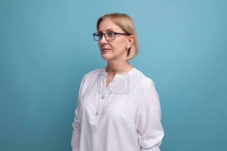 Téléchargez les photos : Portrait d'une femme soignée d'âge moyen aux cheveux blonds à lunettes et chemise blanche. - en image libre de droit