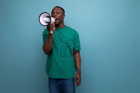 Téléchargez les photos : Un jeune homme africain beau avec une coupe de cheveux courte portant un t-shirt de base parle à l'aide d'un microphone pour attirer l'attention. - en image libre de droit