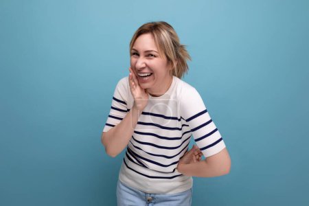 Foto de Foto de cerca de una adorable chica rubia feliz alegre en un suéter casual a rayas sobre un fondo azul con espacio para copiar. - Imagen libre de derechos