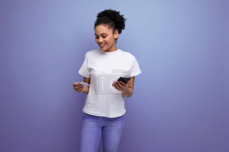 Téléchargez les photos : Jeune femme latine charmante vêtue d'un t-shirt blanc tient une carte en plastique et un smartphone dans ses mains. - en image libre de droit