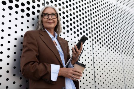 Photo for Successful gray-haired business grandmother dressed stylishly uses a smartphone while walking along the city street. - Royalty Free Image