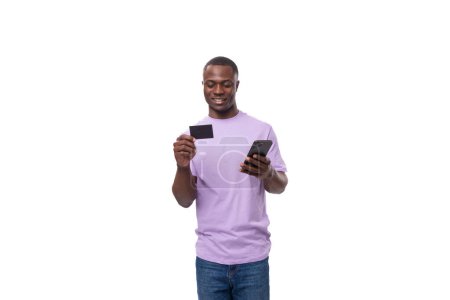 Photo for Young handsome african man in a lilac t-shirt uses a smartphone and a credit card on a white background with copy space. advertising concept. - Royalty Free Image
