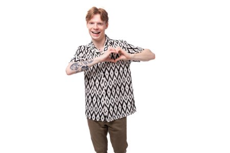 Foto de Un joven amigable con el pelo rojo y un tatuaje en sus brazos está vestido con una camisa de manga corta que muestra un corazón con la mano. - Imagen libre de derechos