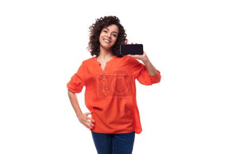 Téléchargez les photos : Jeune dame brune bouclée vêtue d'une élégante chemise orange vif montre l'écran du smartphone. - en image libre de droit