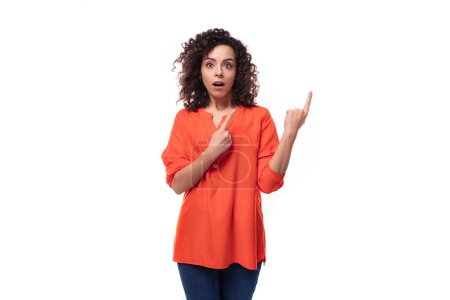 Téléchargez les photos : Jeune femme européenne surprise confus avec les cheveux noirs bouclés en chemise orange sur fond blanc avec espace de copie. - en image libre de droit
