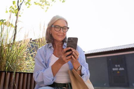 Photo for Mature woman with gray hair and glasses talking on the phone on the street. - Royalty Free Image