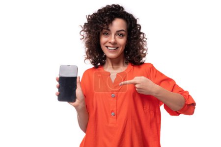 Foto de Joven mujer de negocios caucásica con un peinado rizado vestido con una camisa naranja muestra la pantalla de un teléfono móvil. - Imagen libre de derechos
