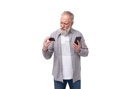 Téléchargez les photos : Un beau grand-père avec une barbe blanche et une moustache, vêtu d'une chemise rayée sur un T-shirt, maîtrise les paiements en ligne à l'aide d'un smartphone et d'une carte plastique. - en image libre de droit