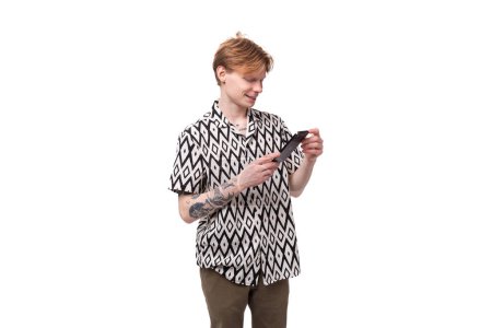 Téléchargez les photos : Un jeune homme informel aux cheveux roux et tatoué sur ses bras est habillé d'une chemise à manches courtes avec un motif tapant un message au téléphone. - en image libre de droit