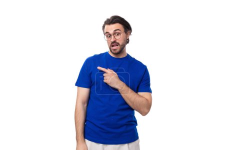 Foto de Joven hombre caucásico inteligente con el pelo oscuro bien cuidado y la barba en una camiseta azul señala su mano a un lado con espacio de copia. - Imagen libre de derechos