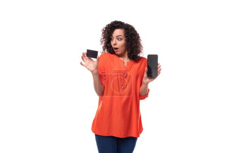 Téléchargez les photos : Jeune femme brune caucasienne aux cheveux bouclés habillée d'un chemisier orange démontre par surprise une carte plastique et un smartphone. - en image libre de droit