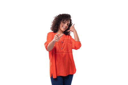 Téléchargez les photos : Jeune femme brune caucasienne frisée élégante vêtue d'un chemisier orange pointe avec son index. concept publicitaire. - en image libre de droit