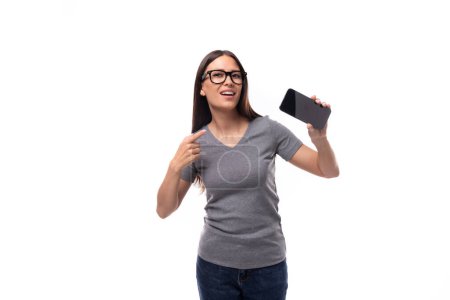 Téléchargez les photos : Jeune promotrice femme en lunettes vêtue d'un t-shirt gris utilise un smartphone sur fond blanc avec espace de copie. - en image libre de droit