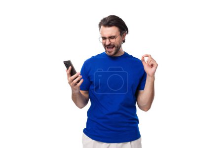 Téléchargez les photos : Jeune homme brune bien entretenu avec une barbe dans un T-shirt regarde une vidéo sur un smartphone. - en image libre de droit