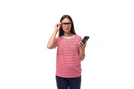 Téléchargez les photos : Charmante jeune femme caucasienne aux cheveux raides avec des lunettes et dans un t-shirt rayé tient un smartphone dans sa main. concept de style de vie. - en image libre de droit