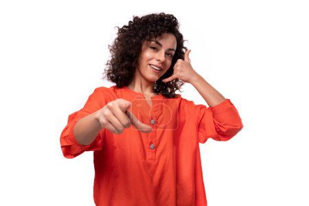 Téléchargez les photos : Jeune dame caucasienne aux cheveux bouclés noirs habillé d'un chemisier orange pointe son doigt vers la caméra. - en image libre de droit
