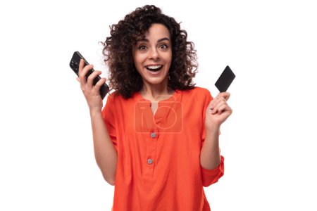 Téléchargez les photos : Une jeune femme européenne aux cheveux noirs bouclés tient une carte plastique et un téléphone dans ses mains et sourit. - en image libre de droit