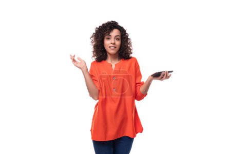 Téléchargez les photos : Jeune femme brune charmante vêtue d'un chemisier orange tient un smartphone dans sa main. - en image libre de droit