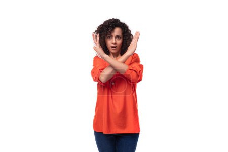 Téléchargez les photos : Jeune femme européenne aux cheveux noirs bouclés dans un chemisier orange montre les bras croisés. - en image libre de droit