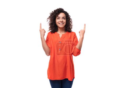 Téléchargez les photos : Jeune dame brune bouclée vêtue d'une élégante chemise orange vif gesticule pointant son doigt vers l'espace de copie. - en image libre de droit